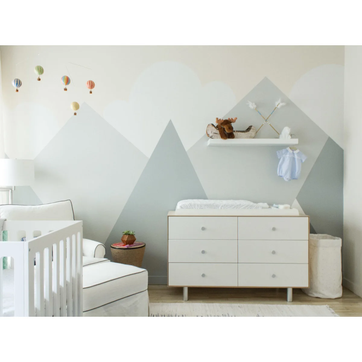 Nursery changing area with a grey mountain mural, white six-drawer dresser and pale upholstered chair