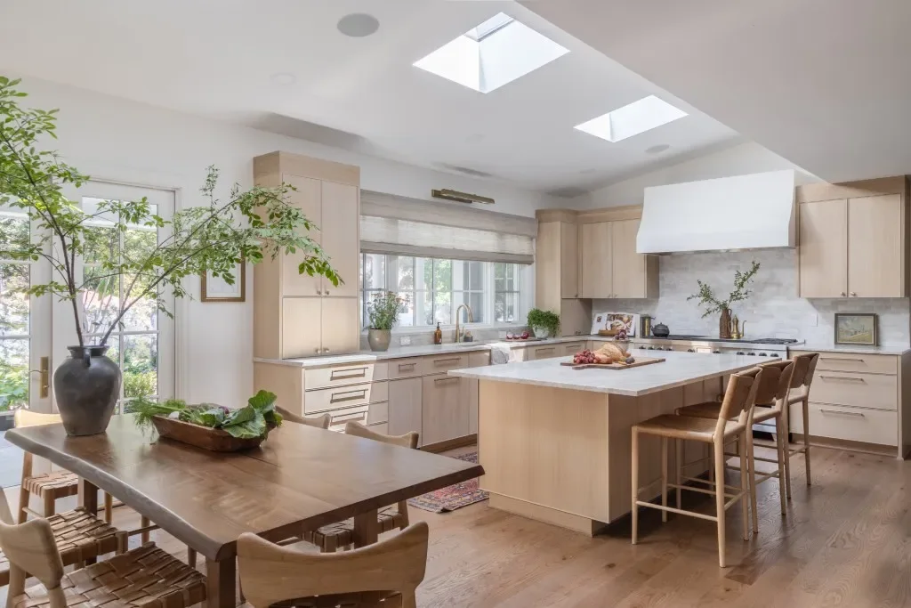 A pale oak kitchen with skylights and a white hood sits behind a darker dining table, woven chairs and a broad island.