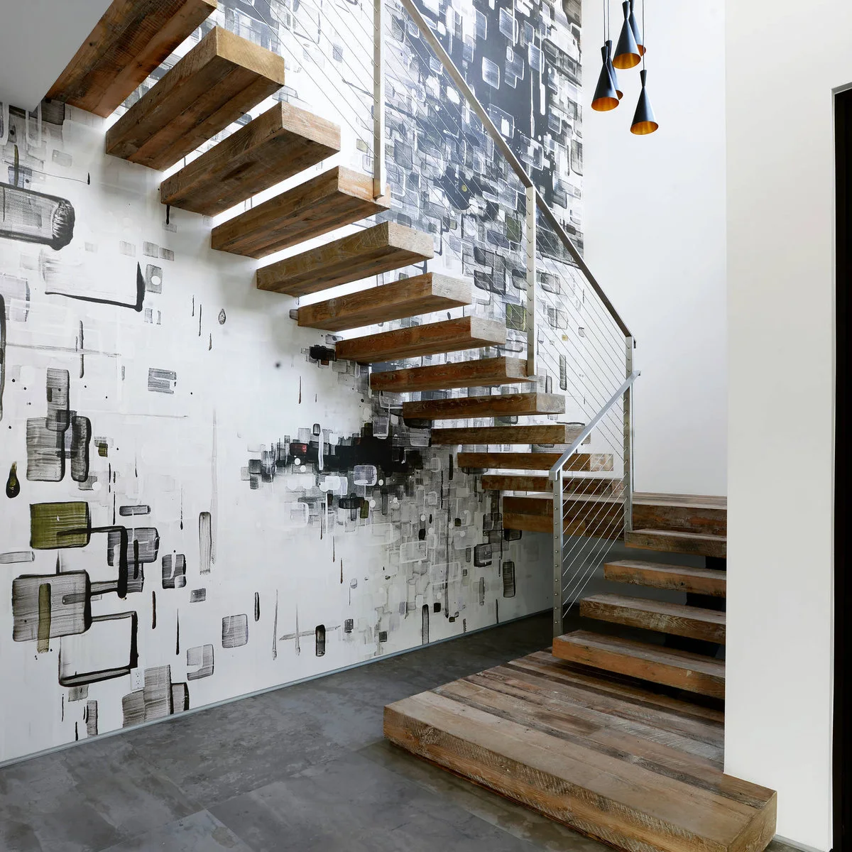 A large black-and-gray graphic wall treatment sits beneath open wood stairs and small pendant lights.