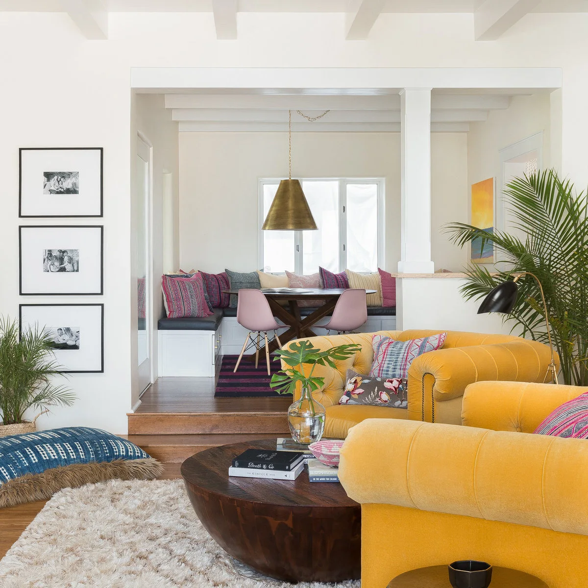 White living area with a yellow rolled-arm sofa, round wood coffee table and colorful dining nook behind
