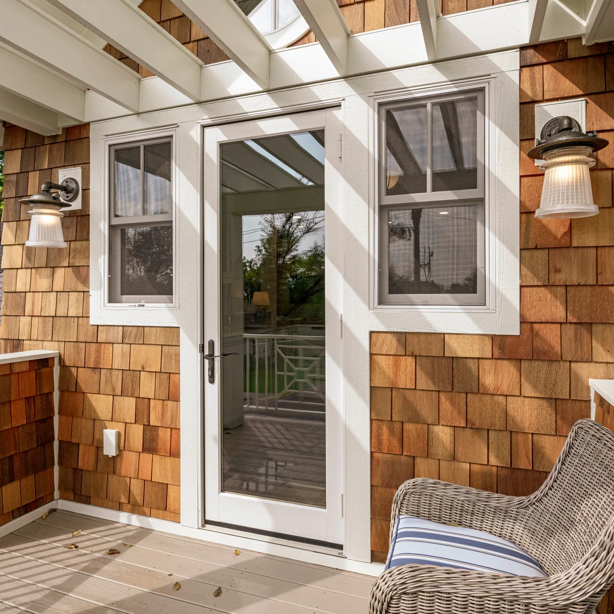 A cushioned woven chair sits to one side of a glass balcony door beneath a pale overhead frame