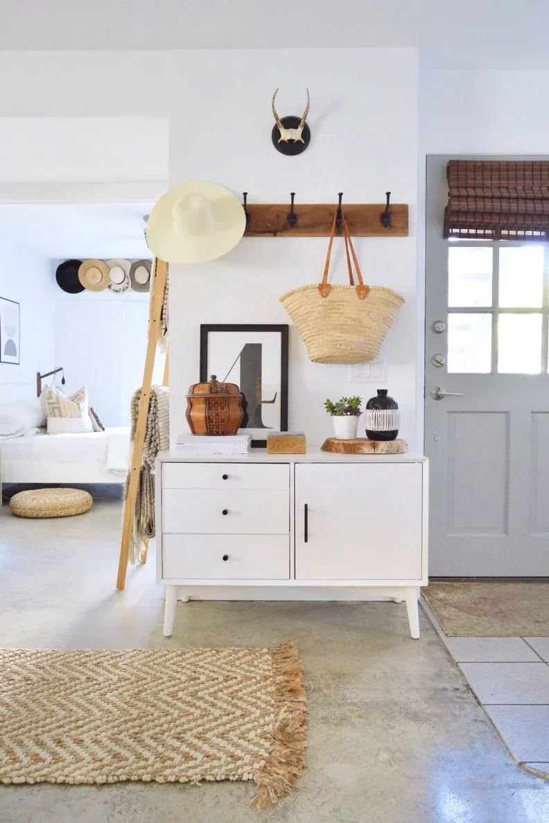 White entry cabinet beneath wooden coat hooks with woven bag, hats, framed picture and light front door.