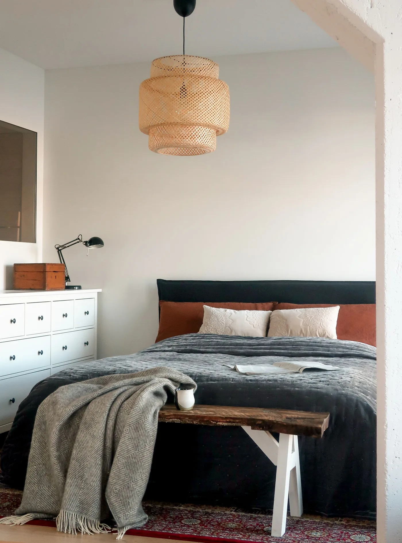 Bedroom with a woven pendant, charcoal bed, rust cushions, white drawer chest and a wood bench on a warm patterned rug