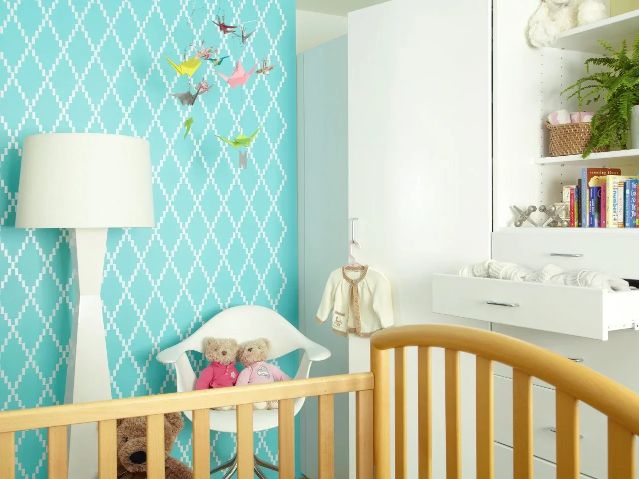 White fitted nursery cupboards include an open pull-out surface beneath shelves beside a turquoise patterned wall.