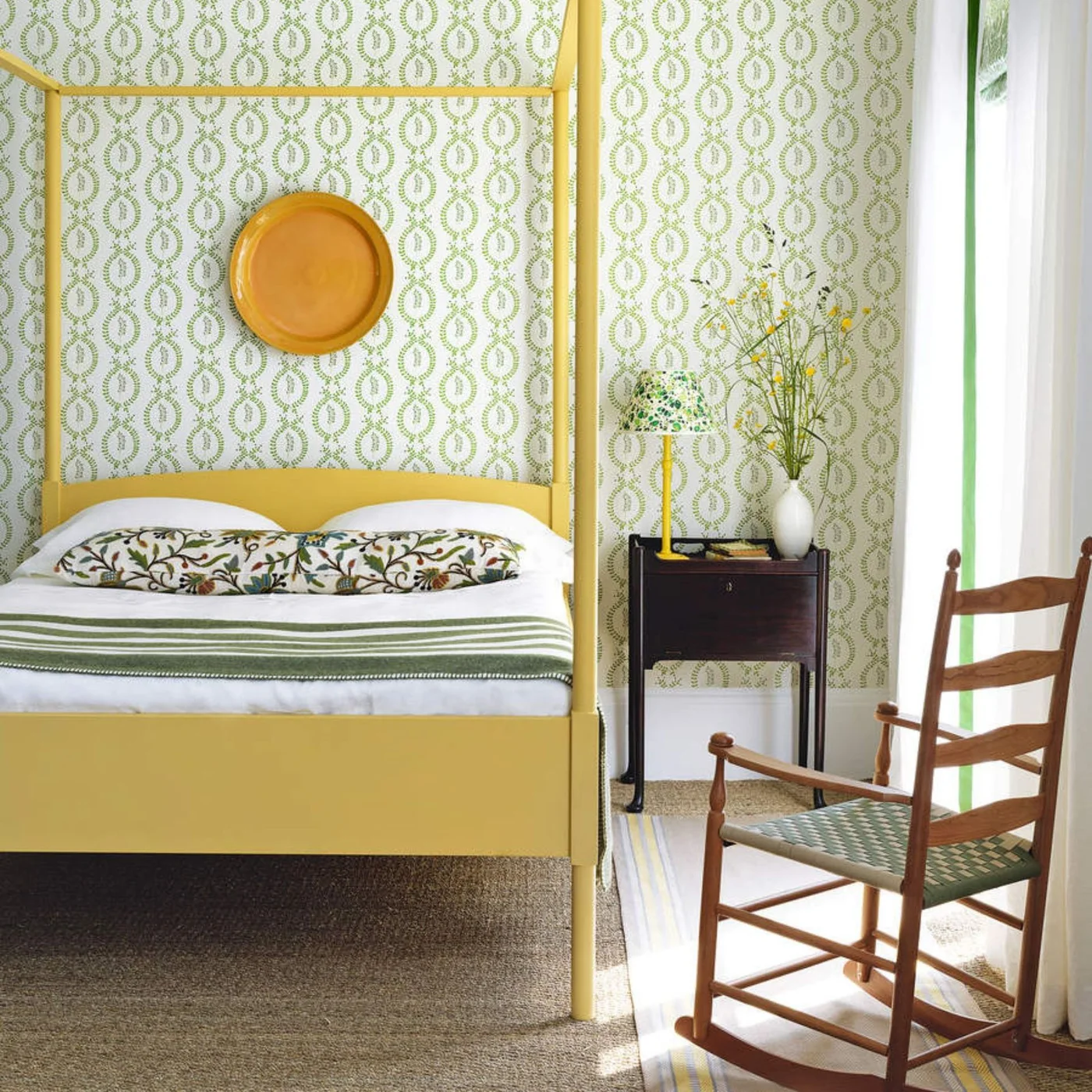 A yellow four-poster bed stands against green patterned walls with a striped green throw, botanical cushion and simple wooden chair