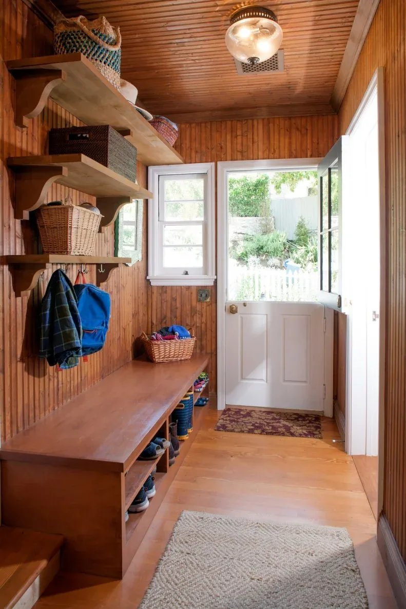 A long wood bench with two levels of shoe shelves runs along a paneled entry beneath coat hooks and basket shelves.