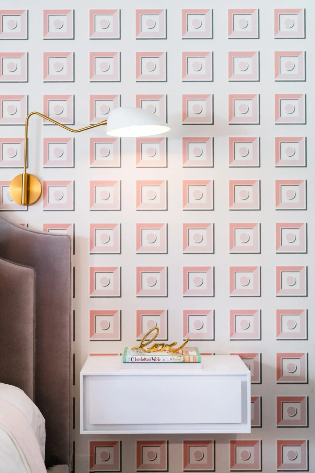 A white-shaded brass-colored wall light sits over a pale bedside drawer against pink geometric wallpaper.