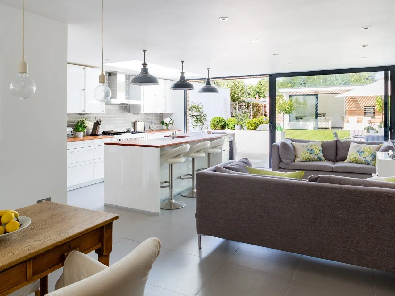 A white kitchen island with pale stools sits beside gray sofas facing a broad garden window.