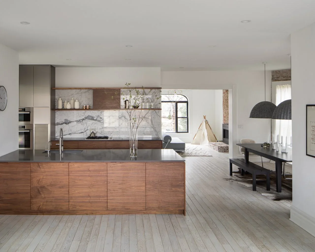 Flat wood island fronts and lower cabinets contrast with dark counters, pale floors and a boldly veined backsplash in an open kitchen.