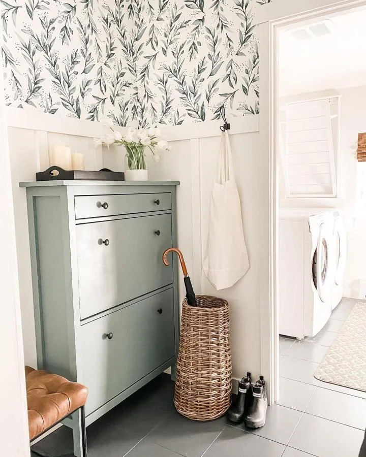Pale green shoe cabinet with dark knobs, a woven umbrella basket and leafy wallpaper beside a laundry doorway.