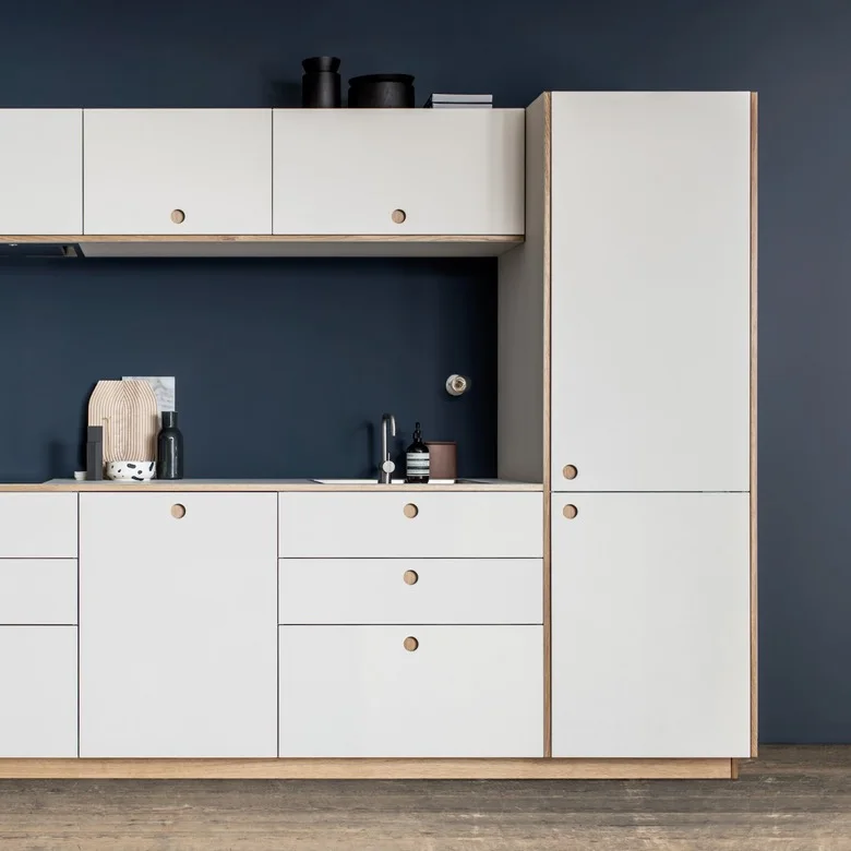 A compact kitchen has flat white cabinets with visible wood-colored edges and circular pulls against a dark blue wall, with a wood-trimmed counter.