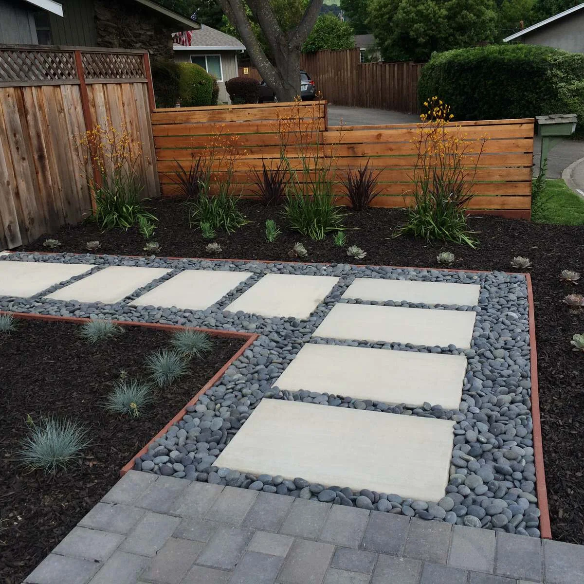 A horizontal wood fence behind a planted bed and pale stepping slabs set in dark river stones.