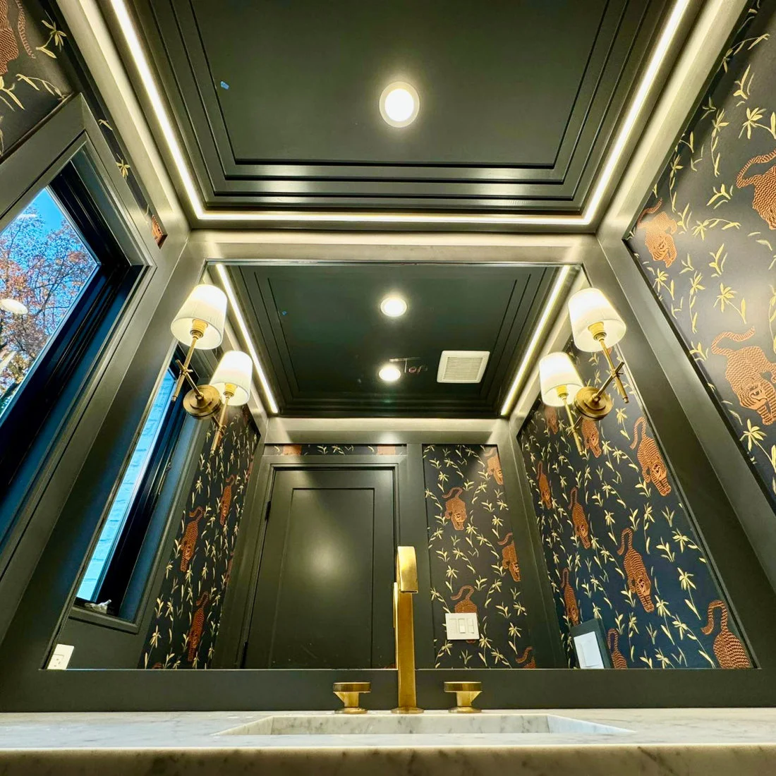 Dark powder-room trim and ceiling panels frame illustrated animal-and-plant wallpaper beside shaded wall lights and a wide sink mirror