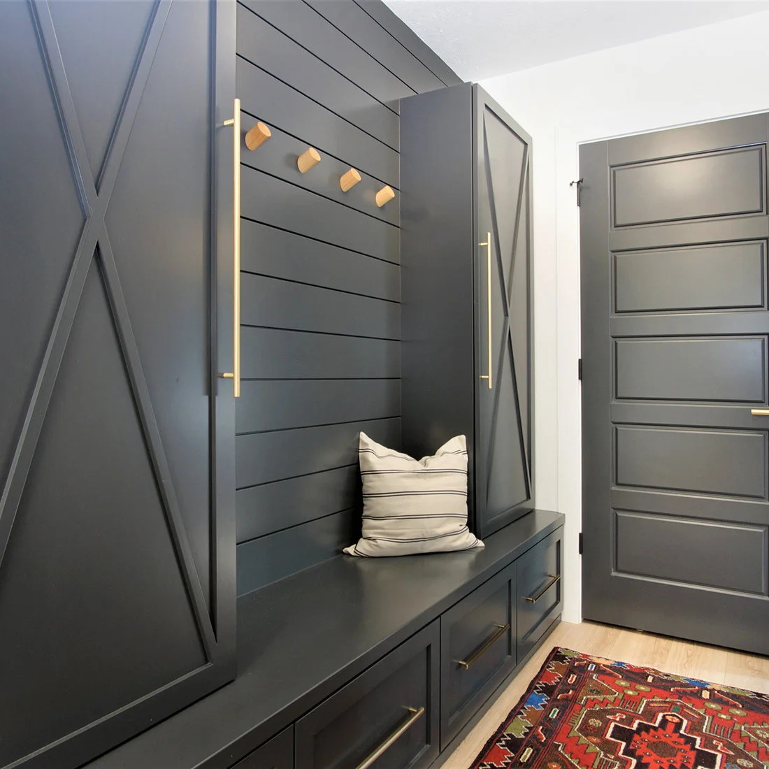Dark blue-gray cupboards and a bench with drawers surround a matching horizontal-board hook wall beside a dark door and patterned rug.