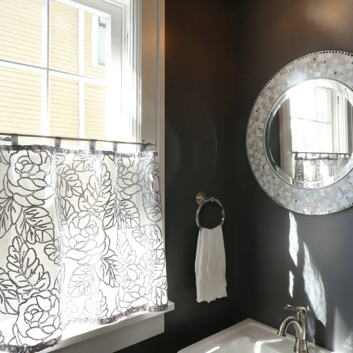 White cafe curtains with a gray leaf outline pattern hang beside a dark wall and a round mosaic-framed mirror.