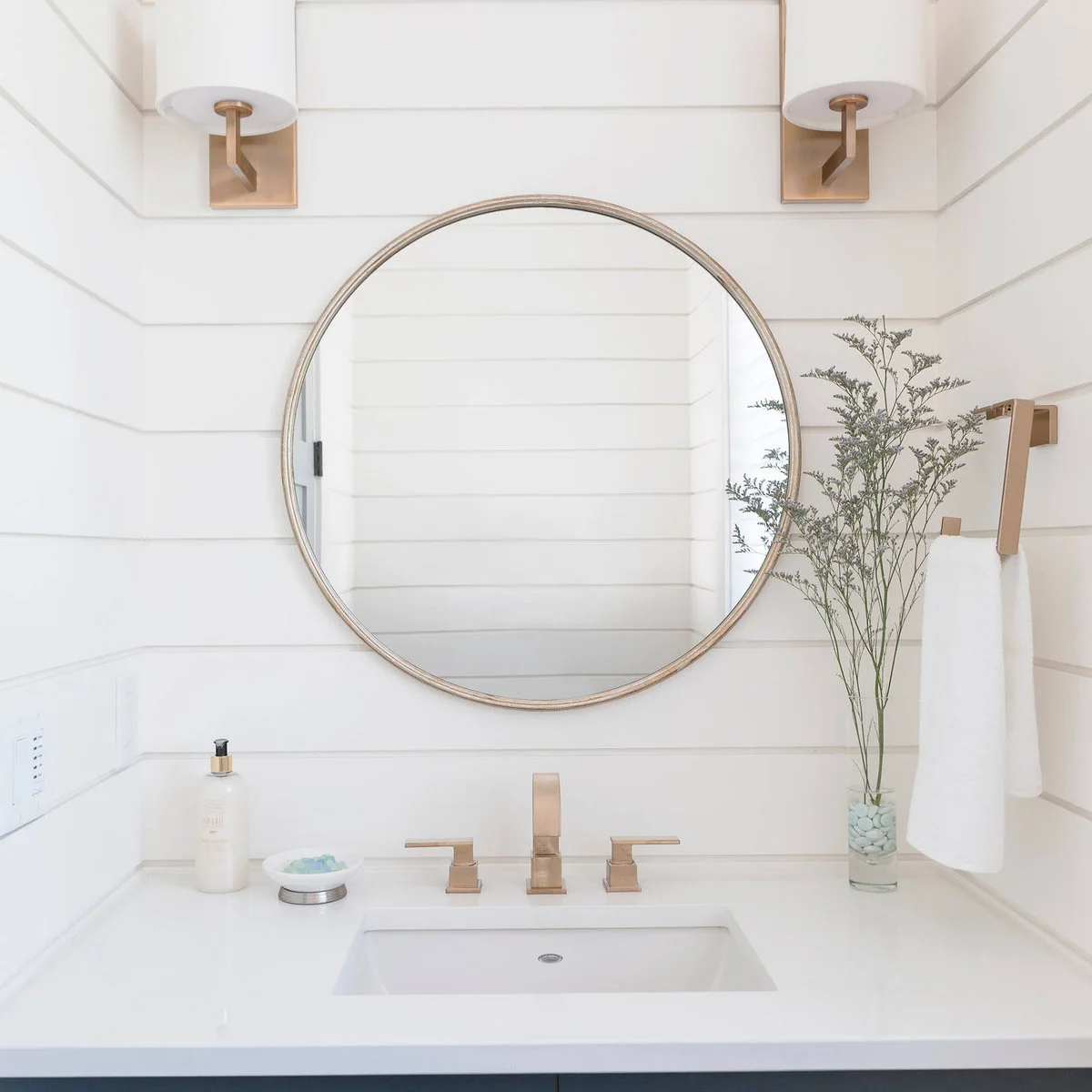 A white towel hangs from a brass-colored rectangular holder beside a round mirror, white paneled walls and square-profile tapware.