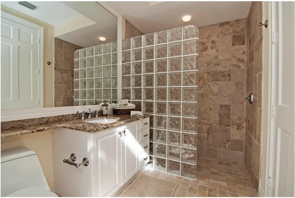 A glass-block partition separates an open shower entrance from a white vanity, with warm brown tiles continuing across the floor.