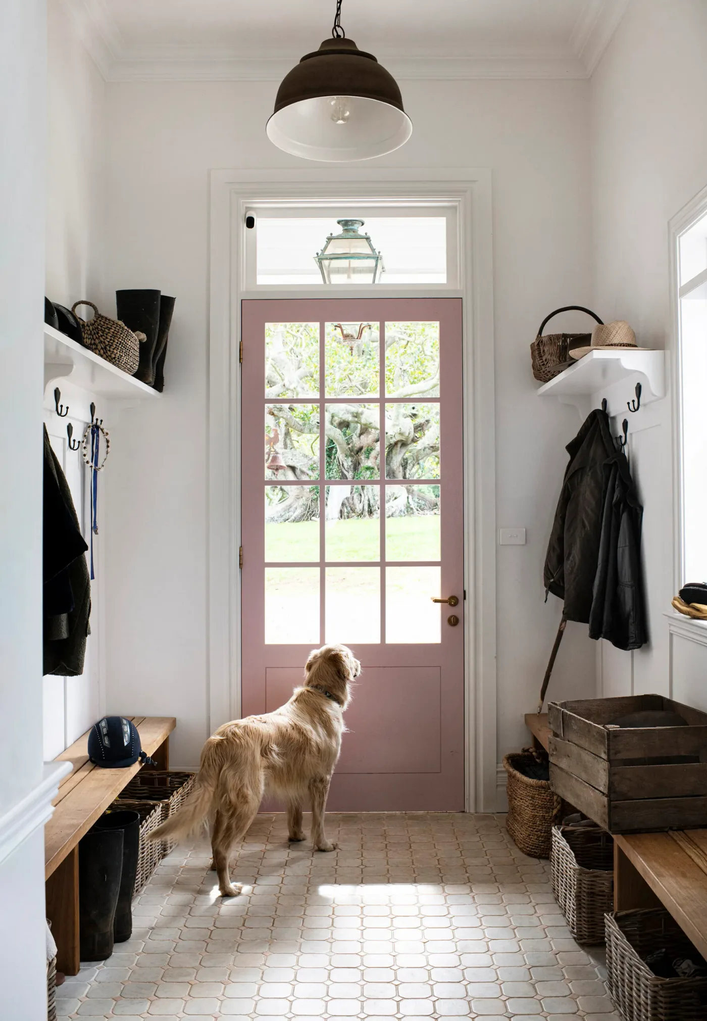 Narrow white entrance with a pink glazed door, shelves and coat hooks on both sides, wooden benches, baskets and tiled floor.