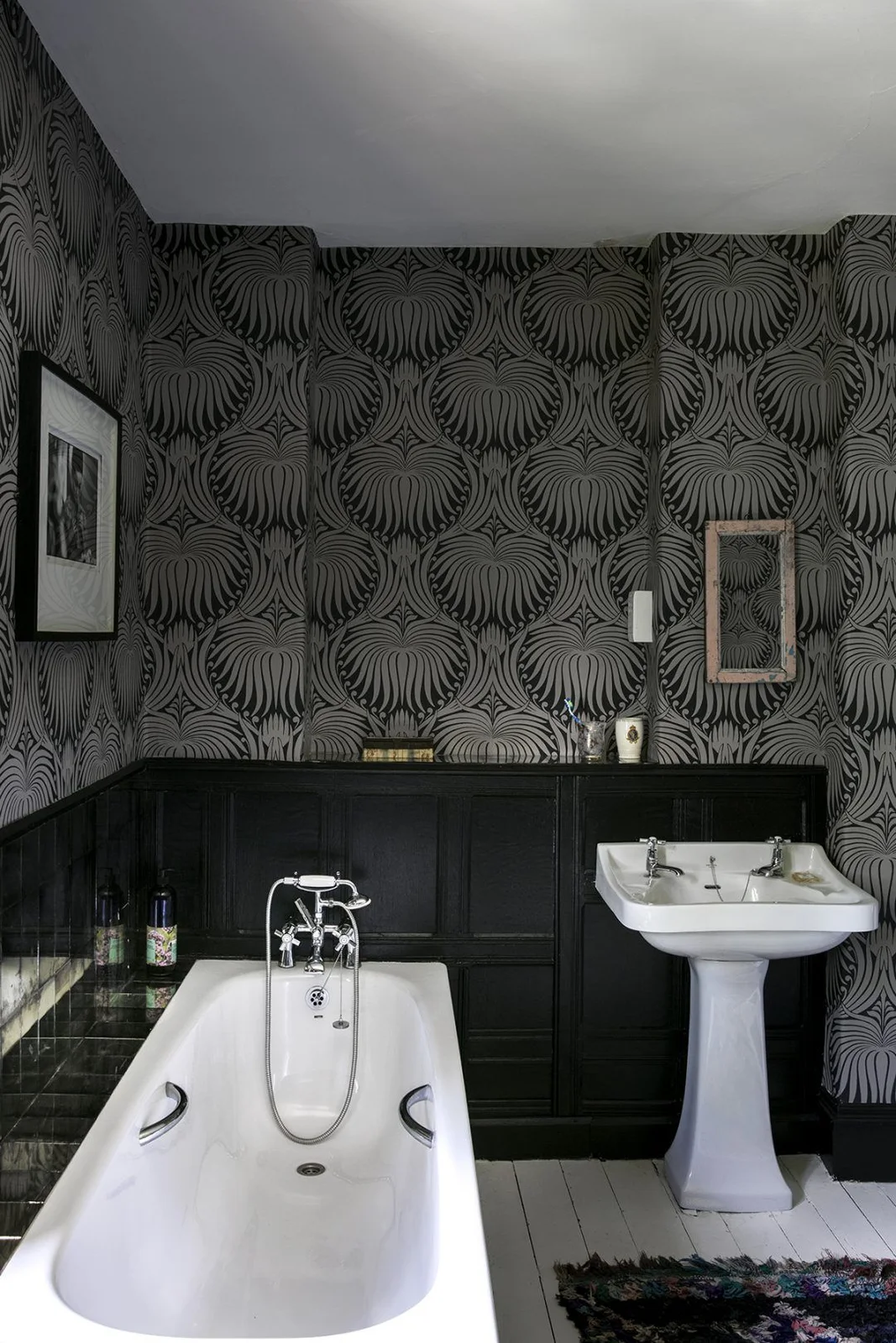A white bathtub and pedestal basin stand below black lower wall paneling and dark leaf-patterned upper walls with framed pictures