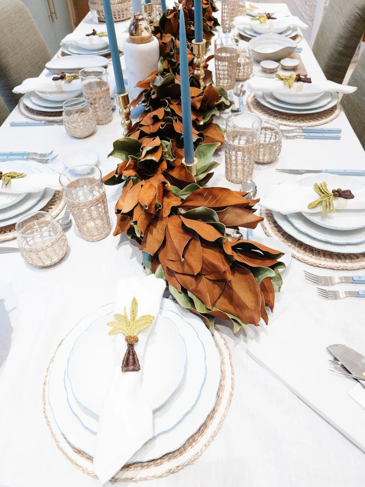A white tablecloth supports blue-edged plates, woven chargers, blue-handled cutlery and blue candles beside a low garland of warm-colored leaves
