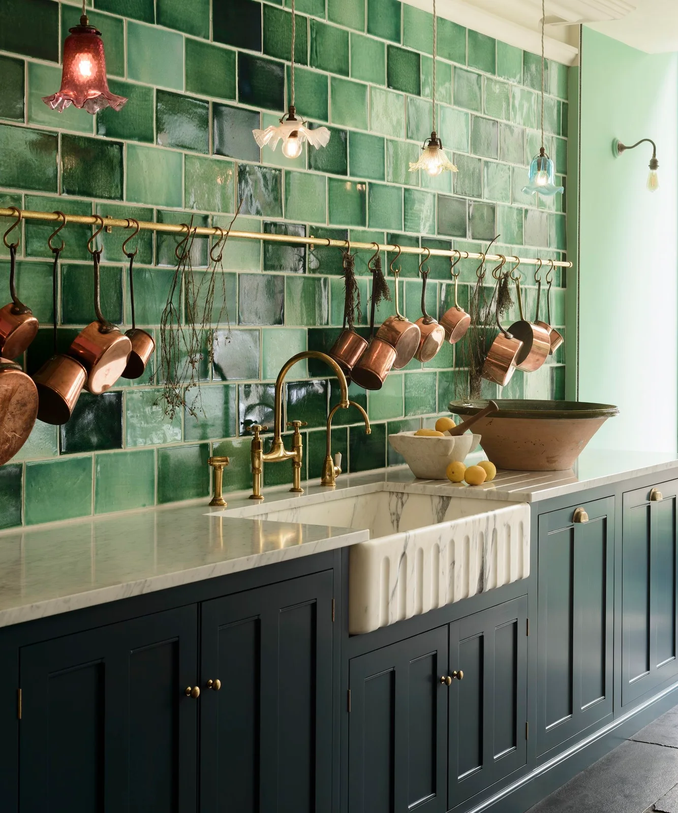 Glossy green rectangular tiles surround a pale fluted sink beneath hanging lights and a rail holding copper-colored pans