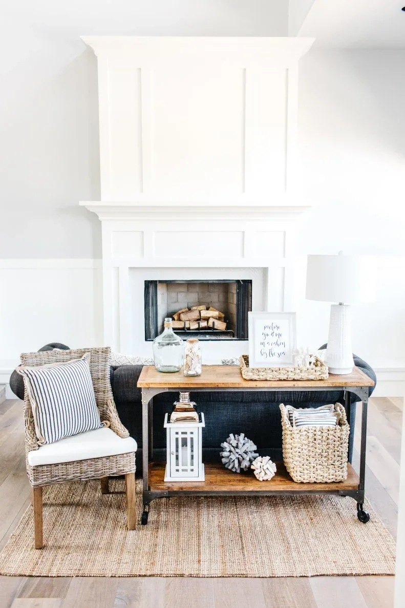 A timber sofa console with a white lamp and green glass bottle holds a woven basket and a white lantern below.