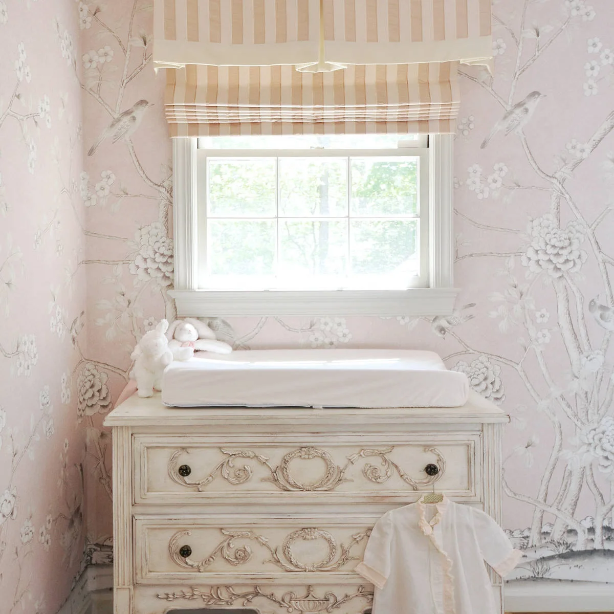 Pale carved nursery chest beneath a window with pink bird wallpaper and a striped blind