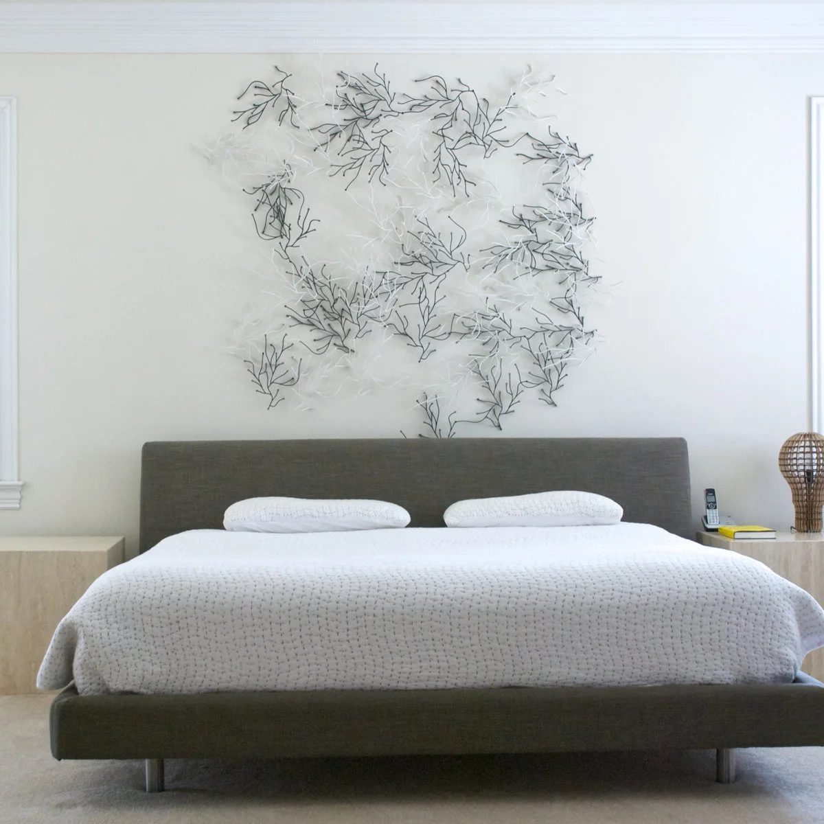 Open white and dark branch-like sculpture above a gray upholstered bed with plain white bedding.