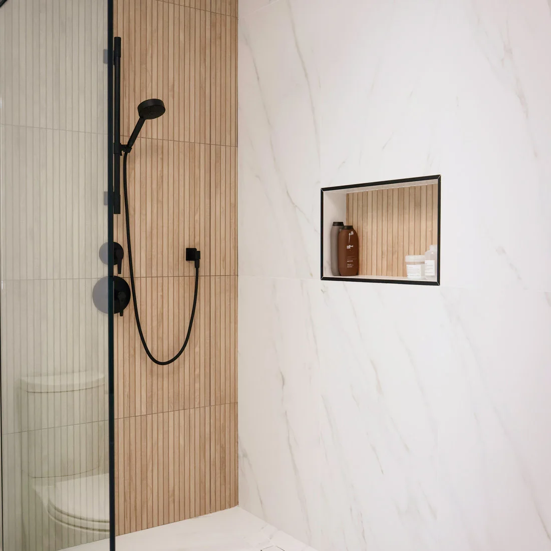 Pale wood-look vertical shower tiles meet a white veined wall with a dark-edged niche and black fittings