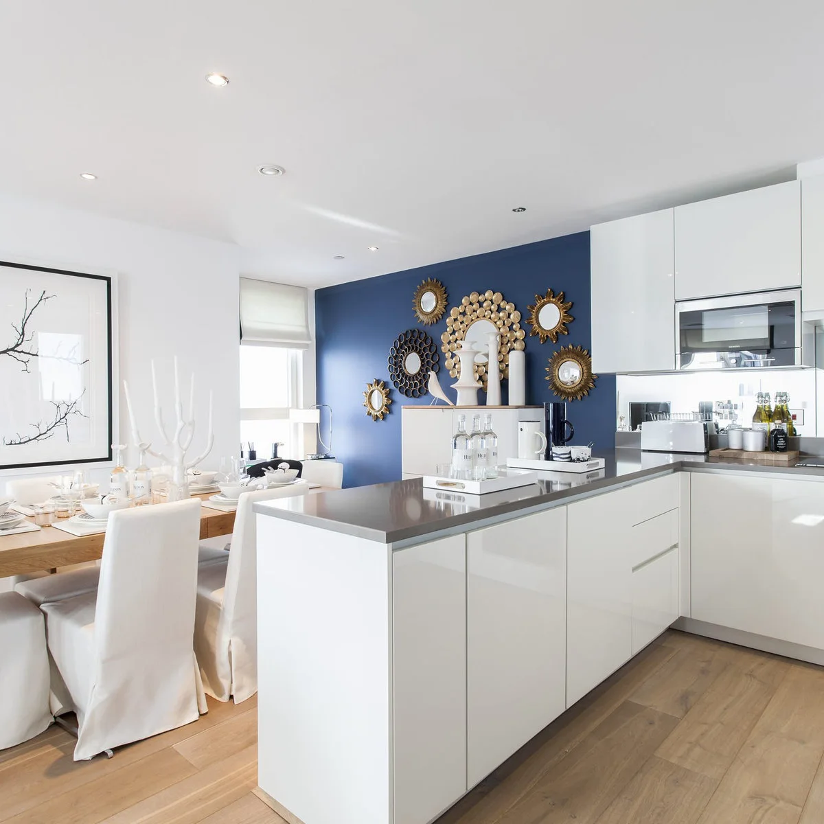 Gold-colored decorative mirrors grouped on a blue wall beside white kitchen cabinets and an eating table.