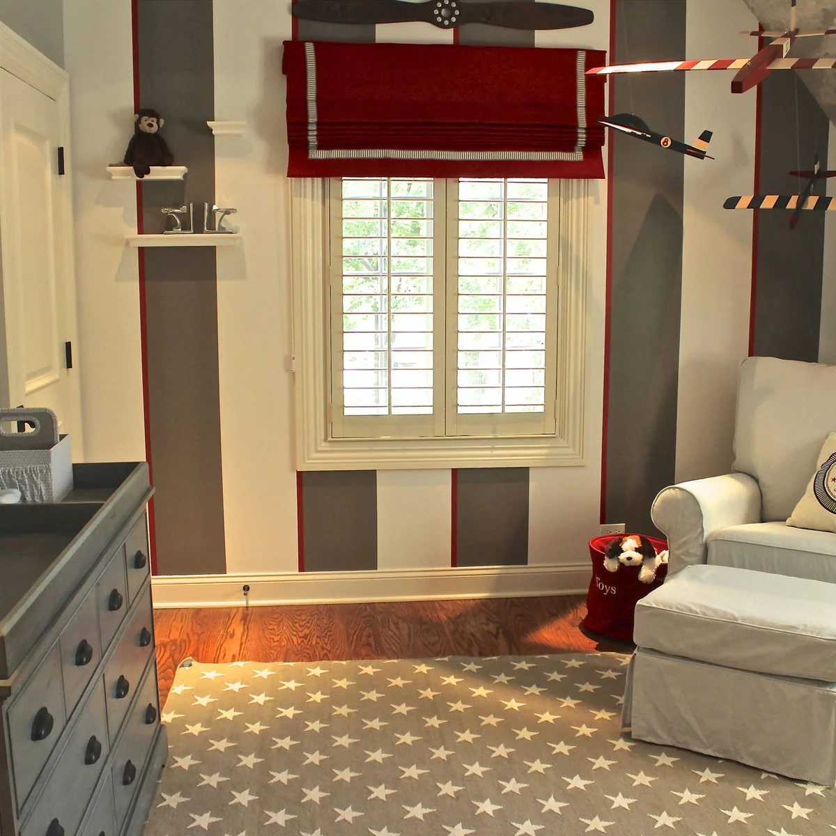 Nursery with broad gray and pale wall stripes, thin red lines, a red shade and a pale chair