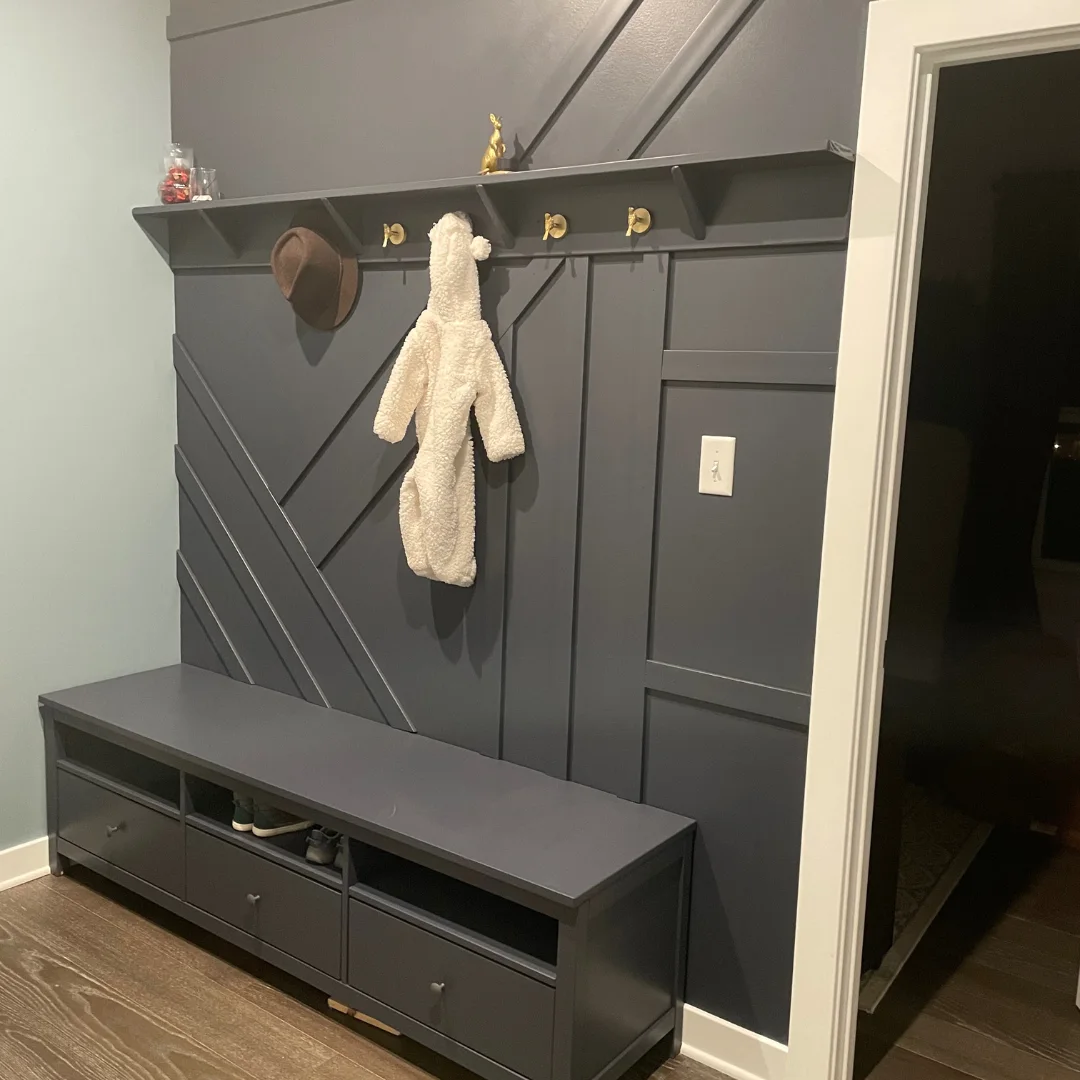 Gray low storage unit beneath a matching gray wall with geometric trim, gold-toned hooks and a shallow shelf.