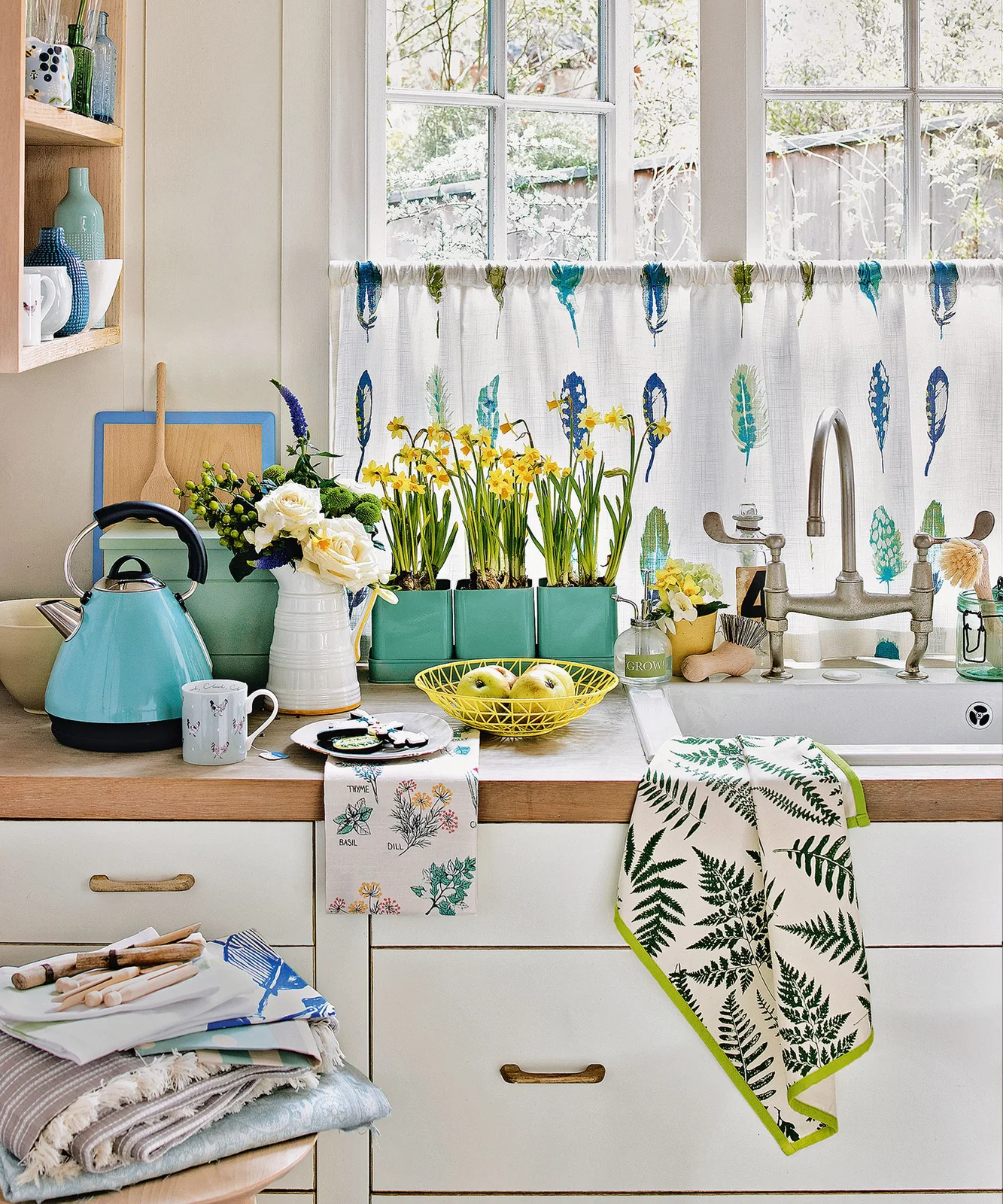 Blue and green feather-pattern café curtain at a kitchen sink with flowers nearby