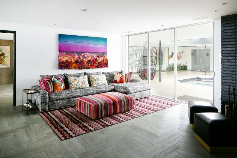Living room with gray sectional, brightly striped ottoman, patterned red-and-cream rug, mixed cushions, colorful landscape artwork and sliding glass doors.