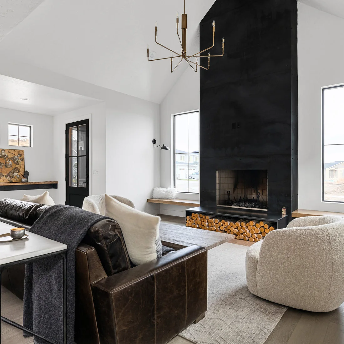 A tall black fireplace column rises beneath a white pitched ceiling, with log storage below, slim wood window benches, pale chairs and a dark brown sofa.