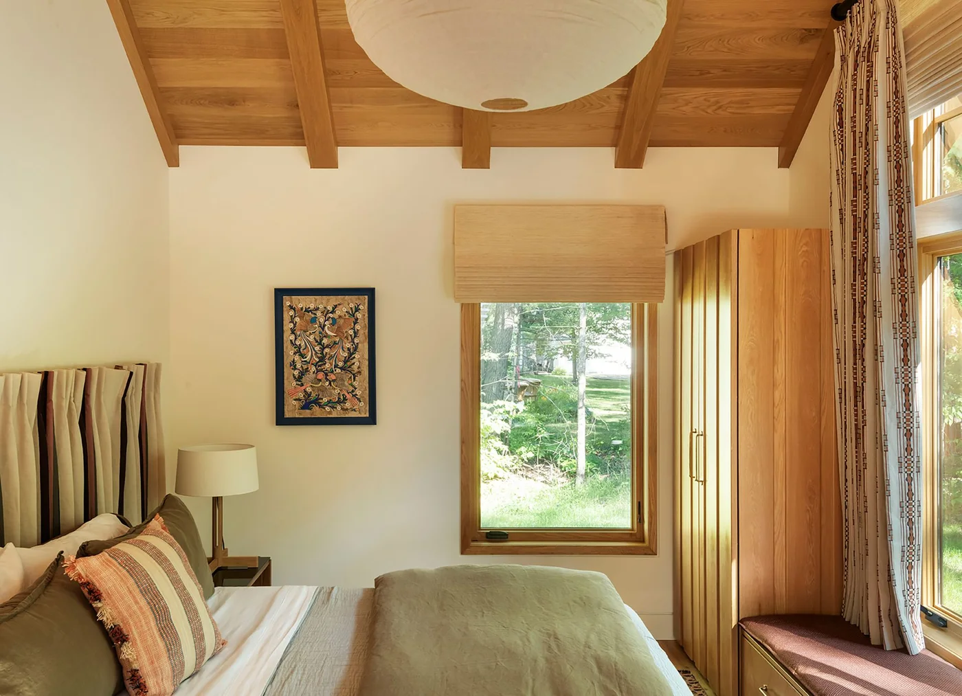 A bedroom has a wood ceiling and beams, a slatted headboard, pale walls, green bedding, a tall wood cupboard and a cushioned seat beside the window.