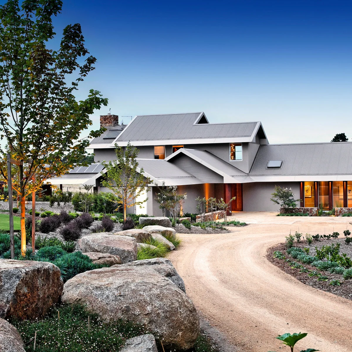 Curving pale driveway beside large rocks, low mixed planting and young trees in front of a contemporary house.