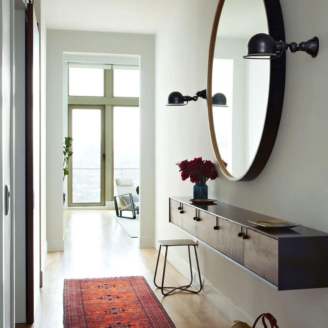 A dark floating drawer cabinet sits beneath a large round mirror and two dark wall lights beside a small stool and red runner.