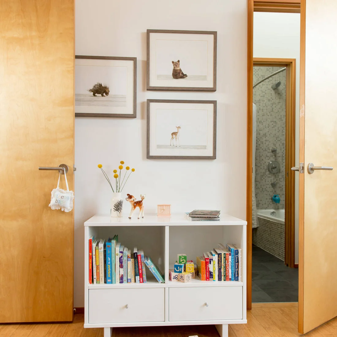 A low white bookcase with drawers displays children’s books and animal pictures beside pale wood doors on a light wood floor.