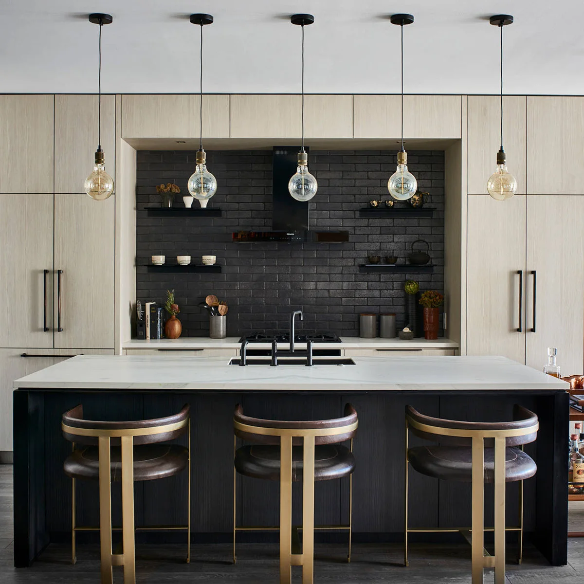 Pale wood cabinets frame a black brick-patterned backsplash and dark island, with pale counters and warm metal stool frames.