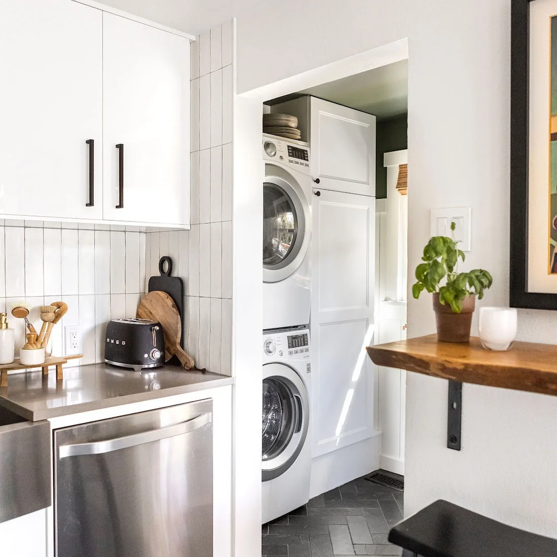 Stacked white laundry machines fit into a white cupboard beside a kitchen with white cabinets, dark handles and a wood shelf.