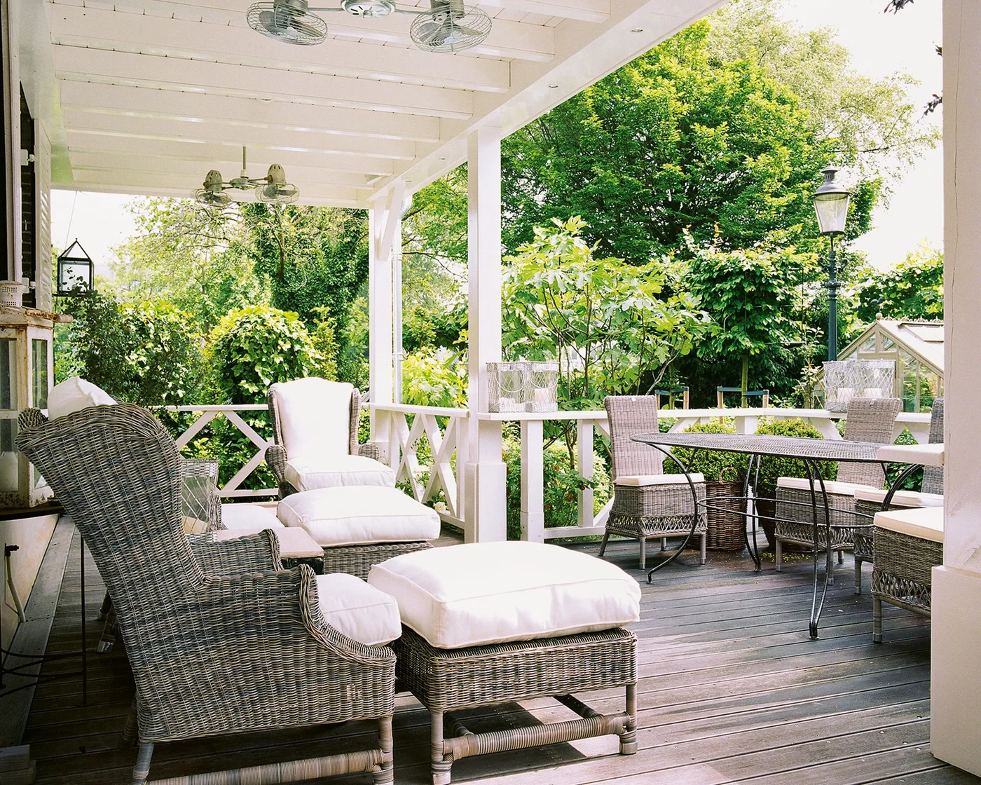 Pale woven seating on a covered white porch looks into a garden with dense shrubs and trees
