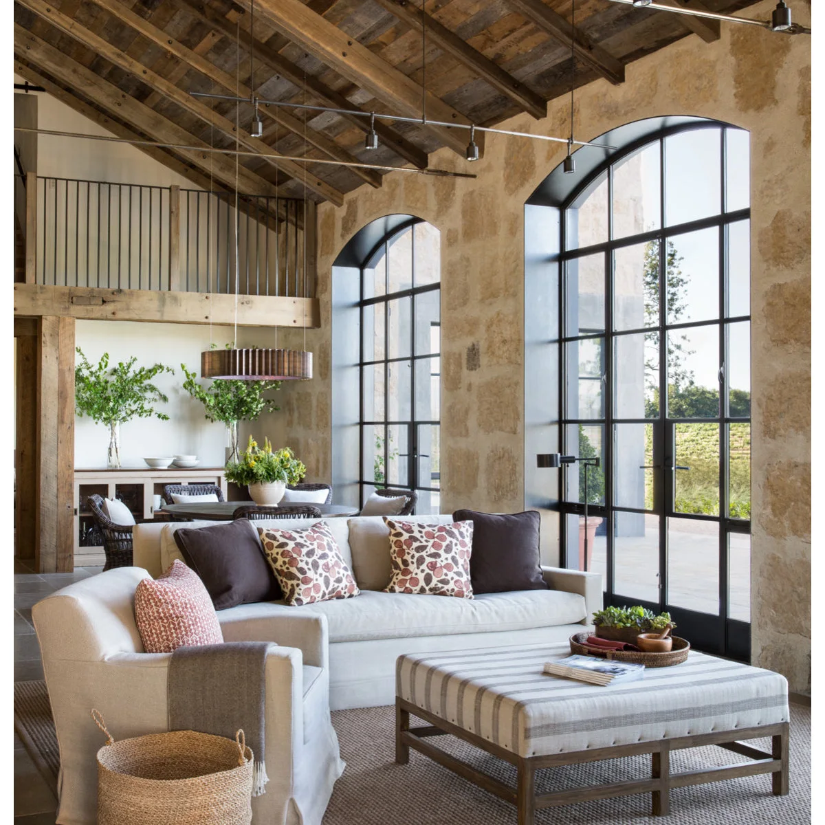 A high living room with exposed wood roof boards, arched black-framed windows, pale seating and a striped upholstered ottoman.
