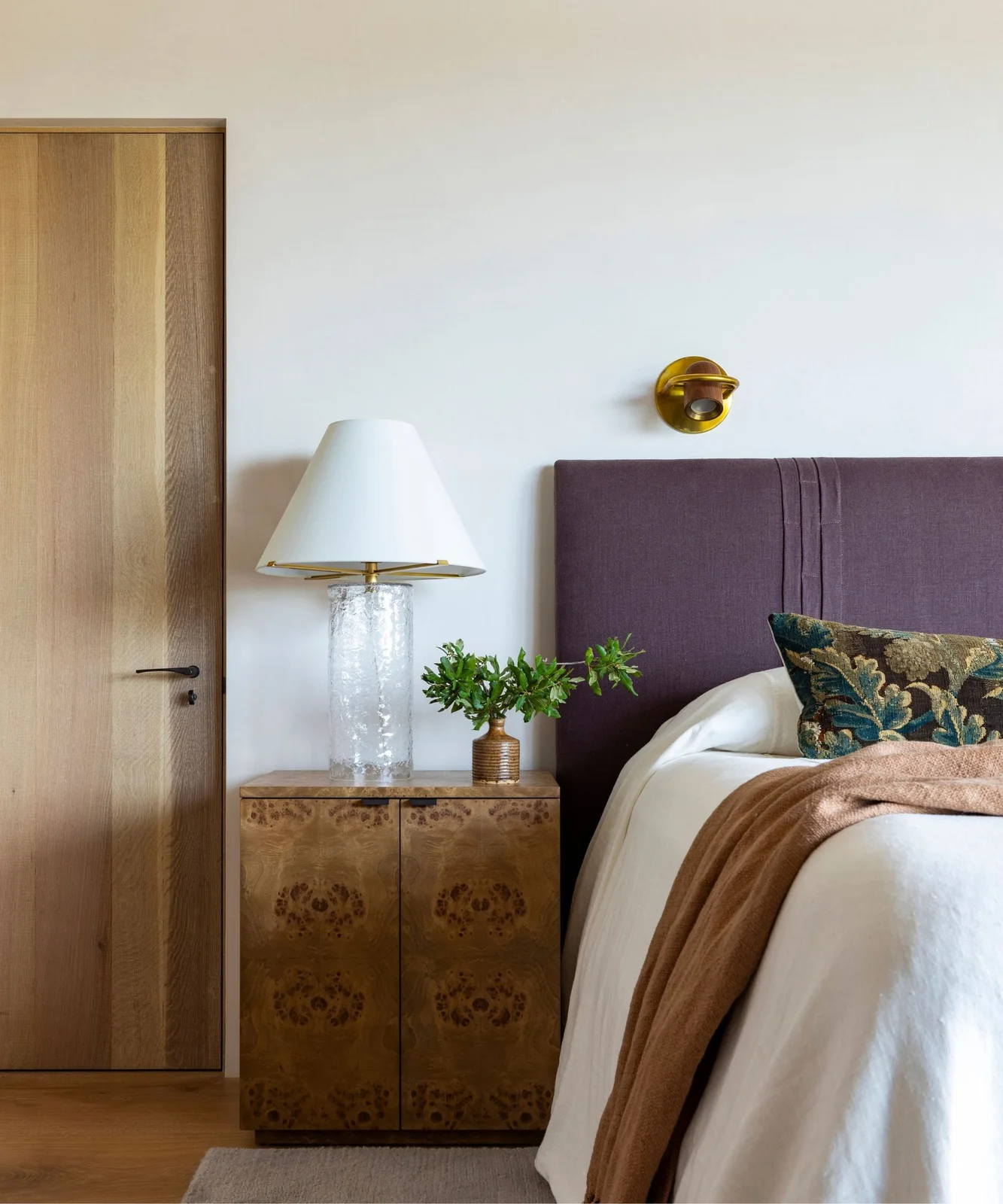 Warm white wall behind a purple headboard with a patterned wooden bedside cabinet and pale bedding.