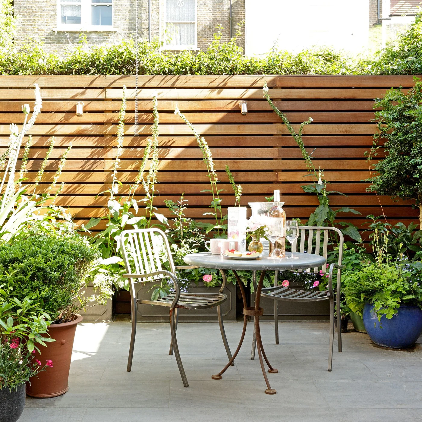 A small round metal table and two chairs sit between potted plants against horizontal timber screening