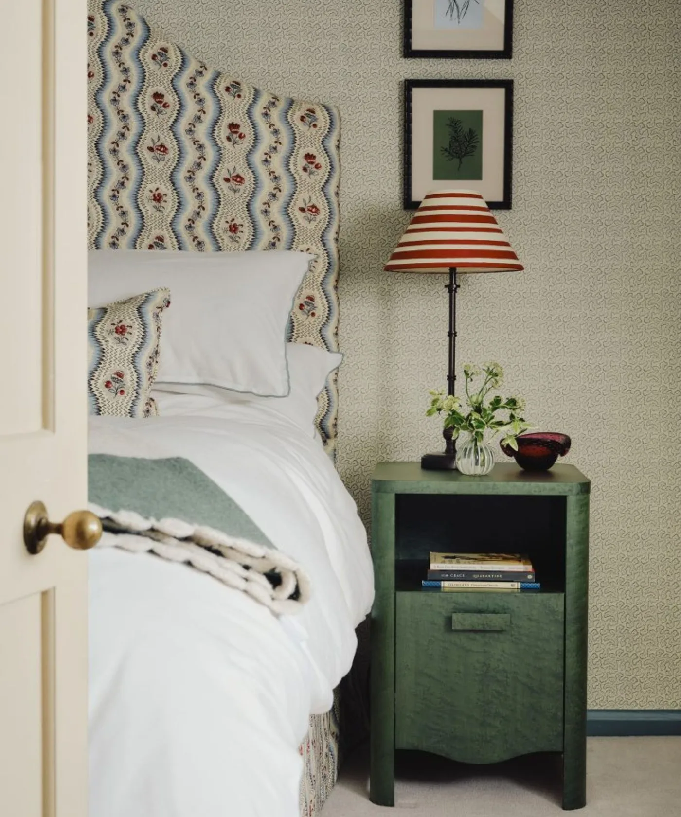 Bedroom detail with tiny pale wallpaper repeat, floral scalloped headboard, green bedside cabinet and striped lamp shade