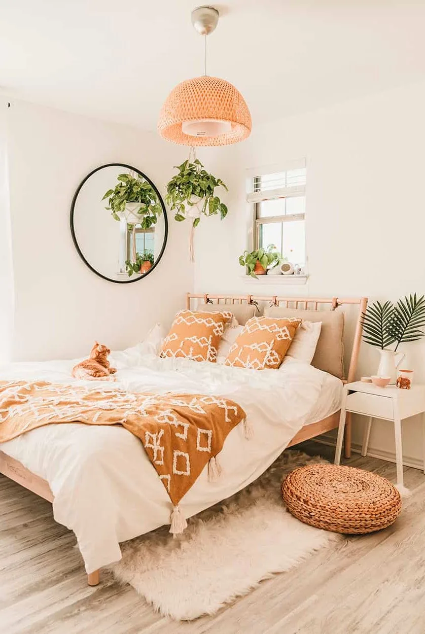 A pale woven bed with white bedding carries orange patterned cushions and a throw beneath a woven shade beside a round mirror and hanging plants