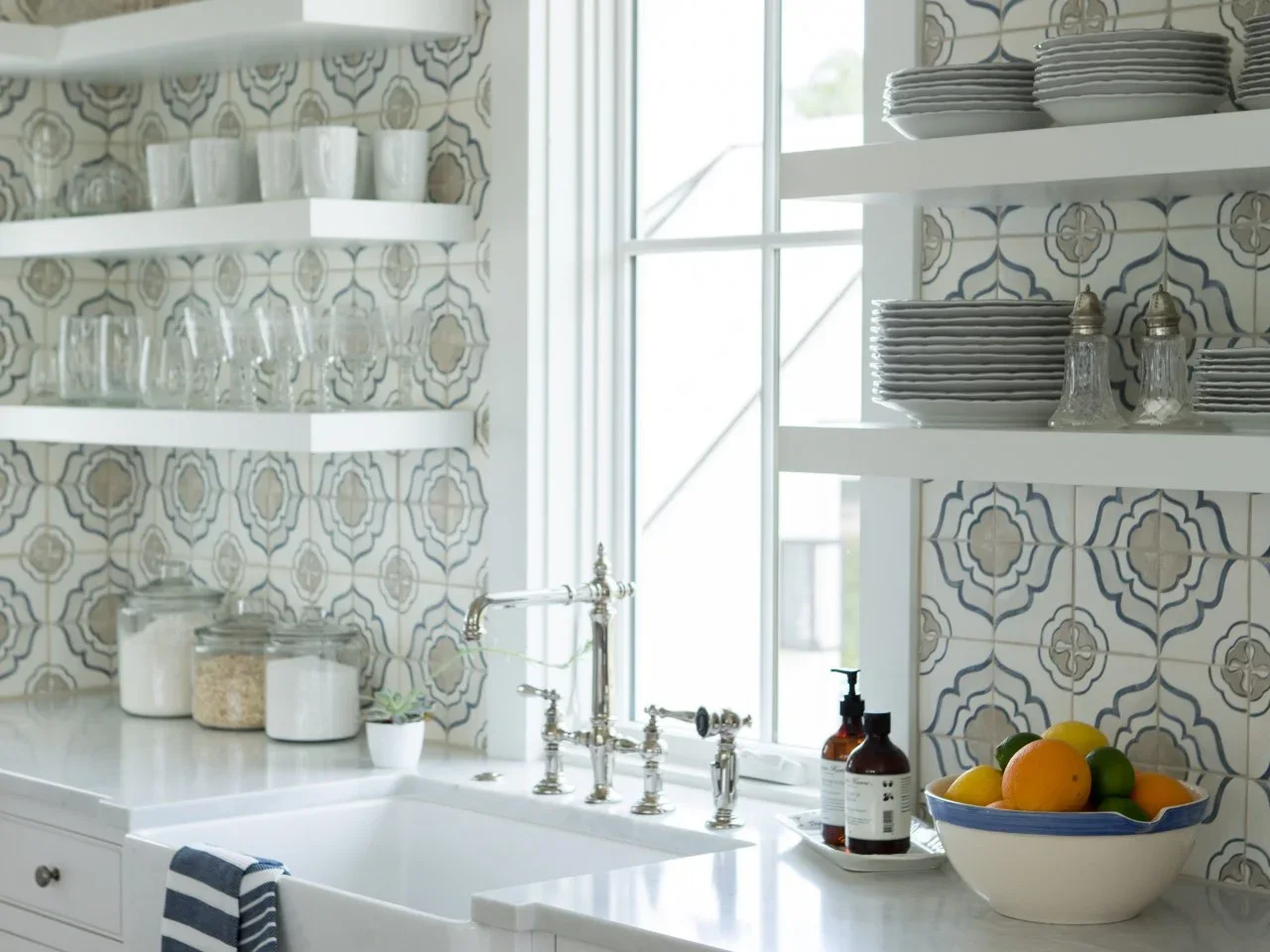 White kitchen tiles with beige and blue floral outlines surround a sink window behind open white shelves.