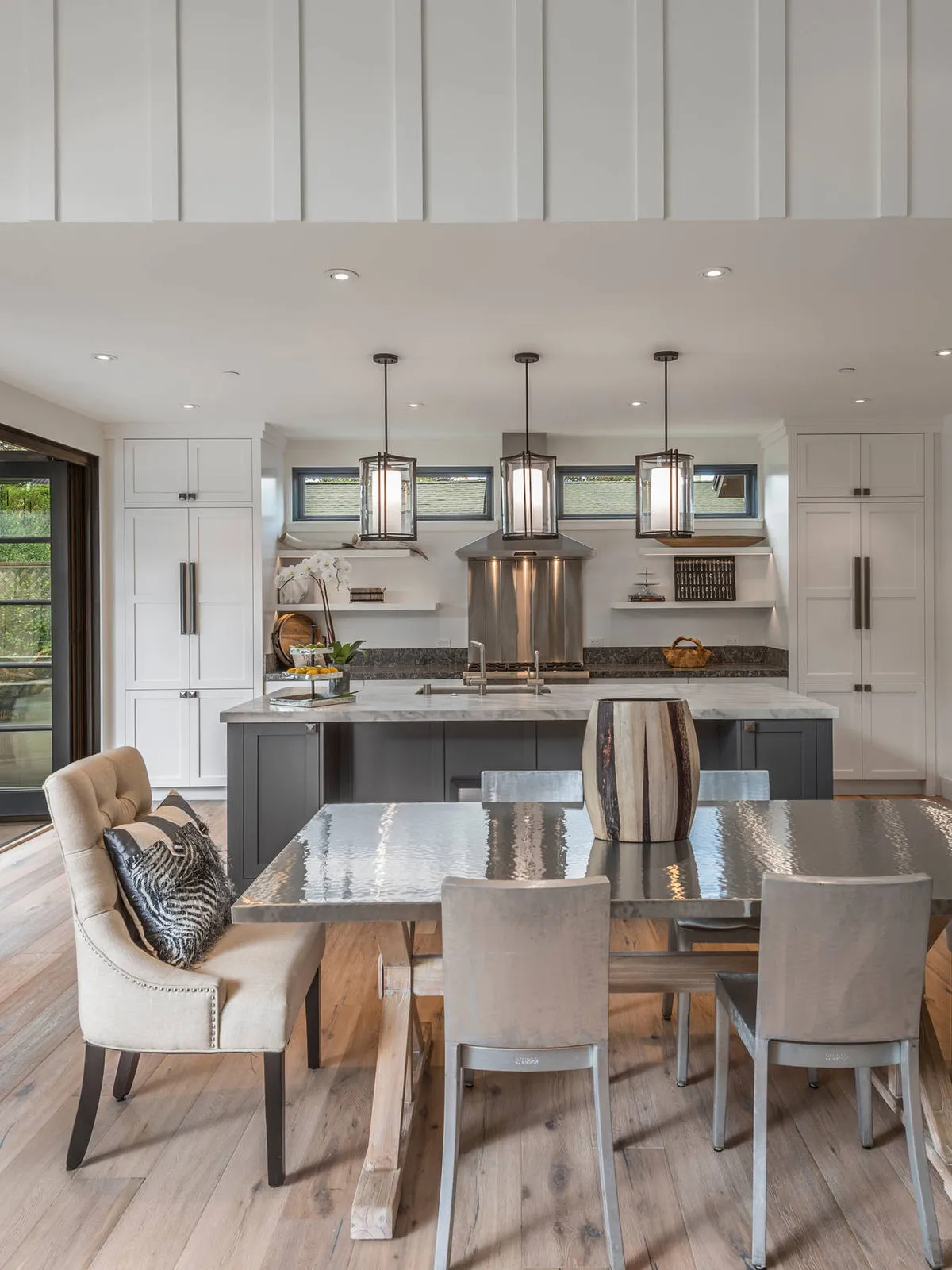 Pale upholstered dining chairs surround a table with a reflective top and timber supports beside a light kitchen.