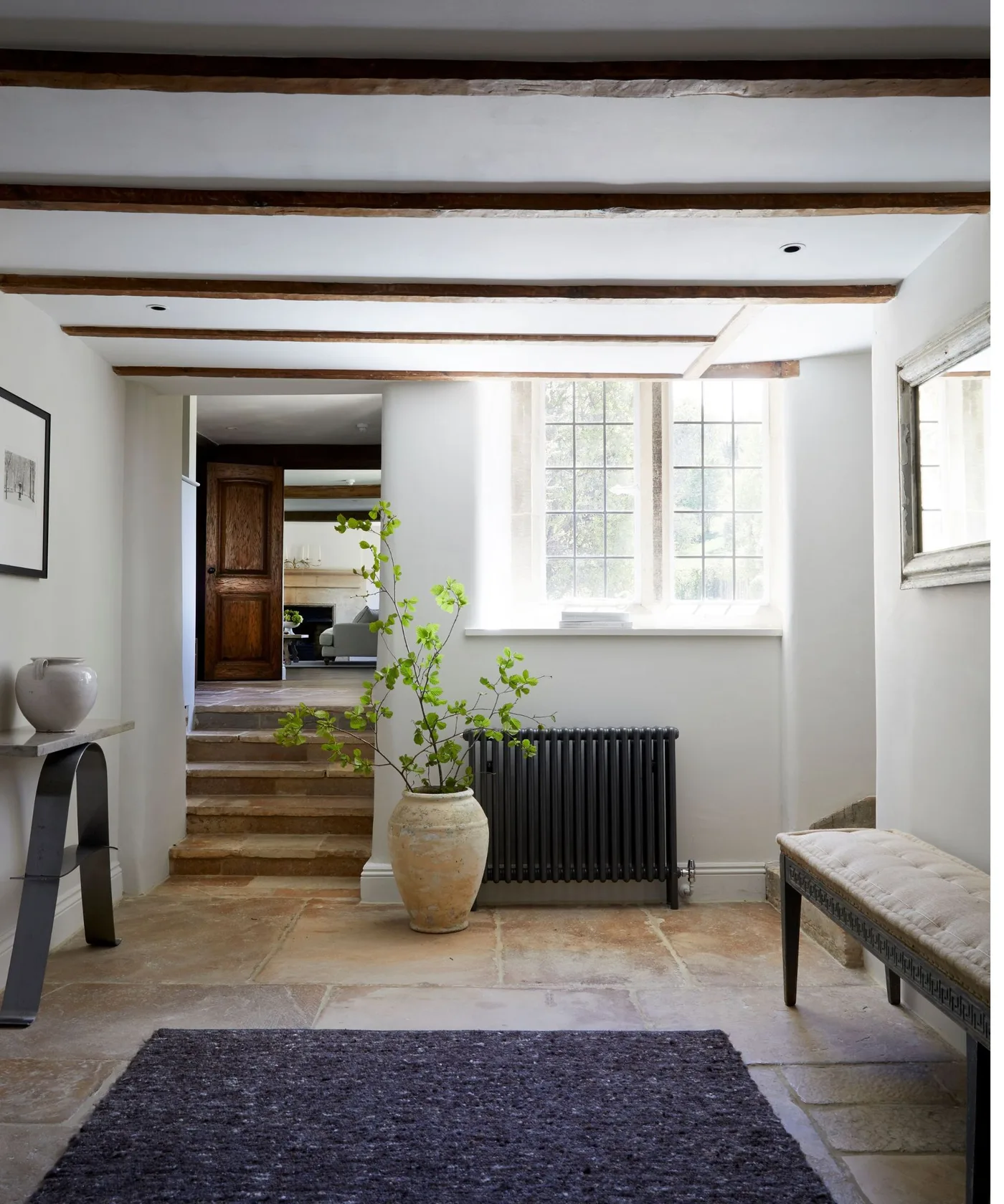 A pale stone floored hall with exposed beams, dark radiator and an upholstered bench